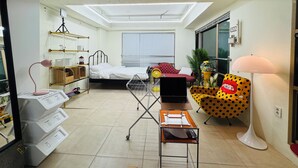 Ein kleines Hotelzimmer mit einem Bett, einem Schreibtisch mit einem Laptop, einem Sitzhocker mit Punkten, einer Stehlampe und einem Fenster mit Jalousien.