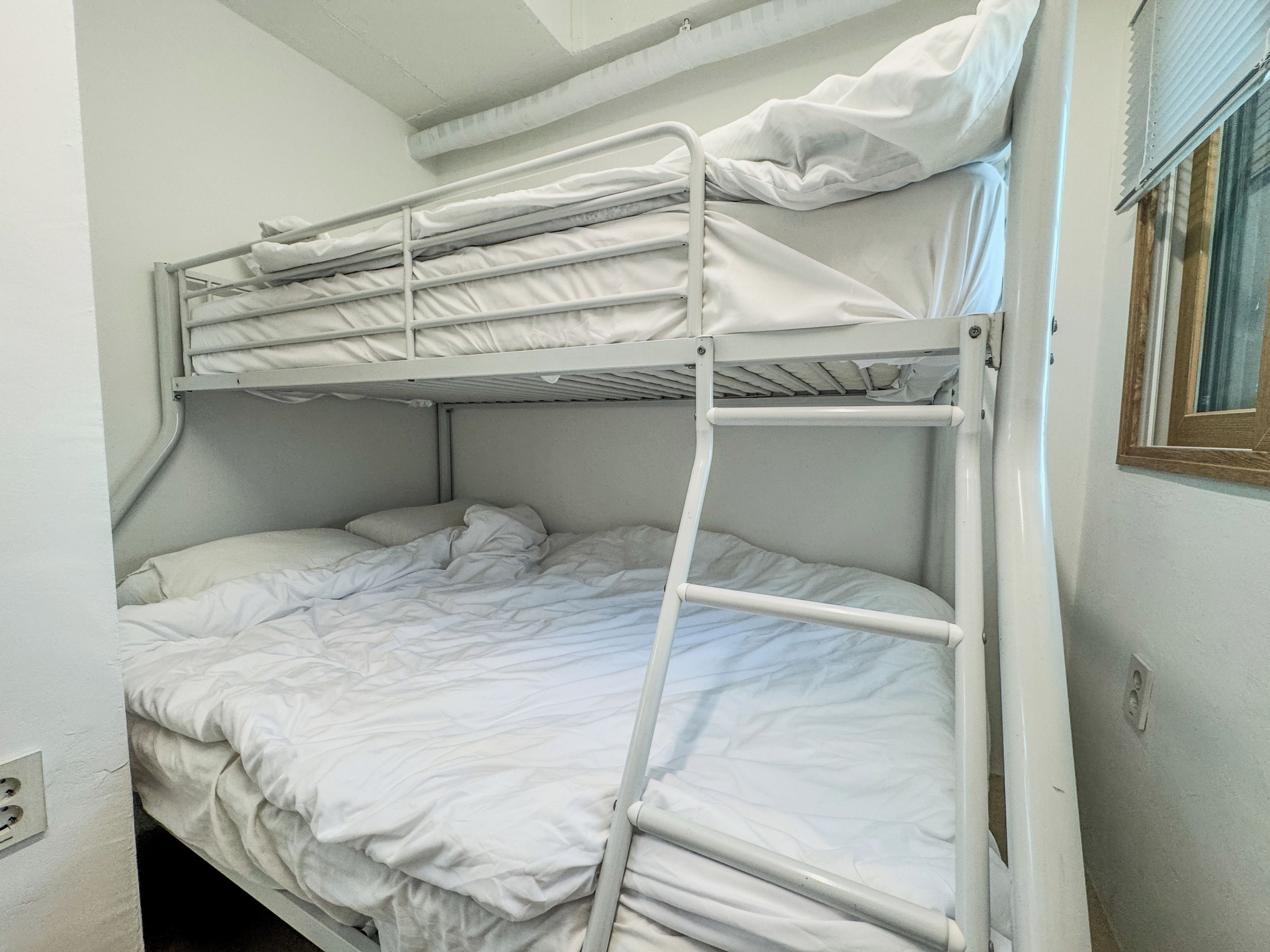 A bunk bed with white bedding, a window with blinds, and a white wall.