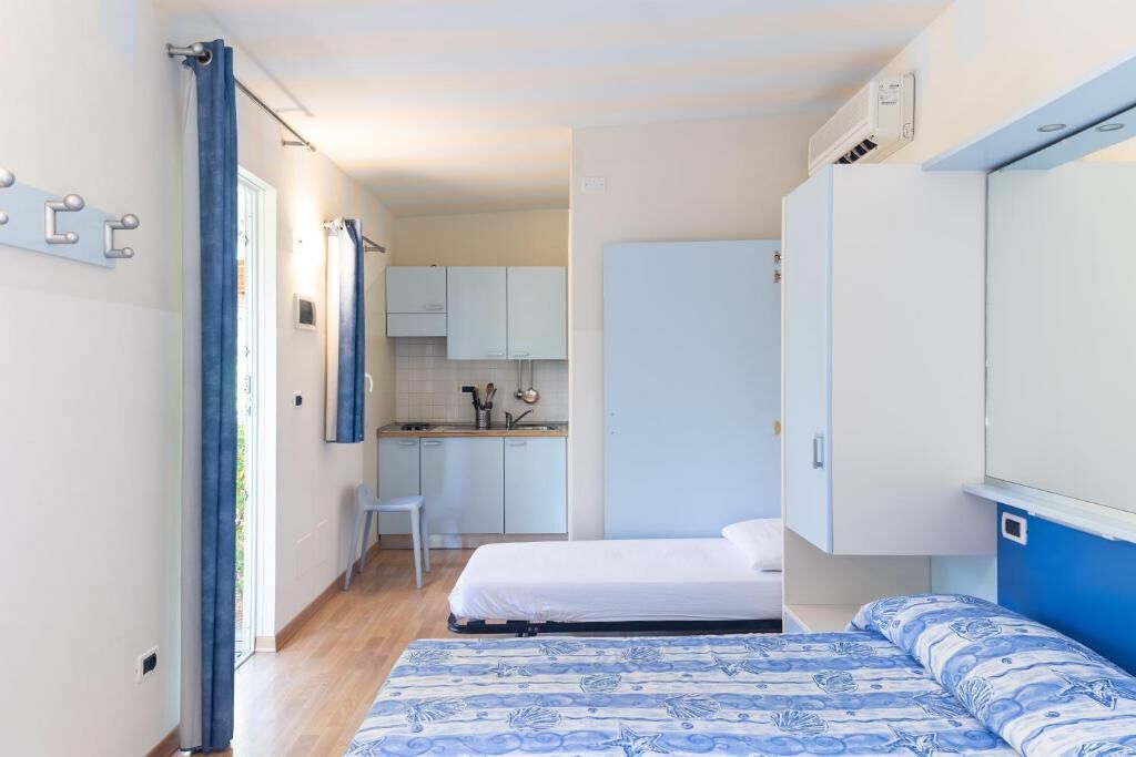 Une chambre à coucher de taille réduite, avec un lit, un petit bureau et, au fond, un coin cuisine.