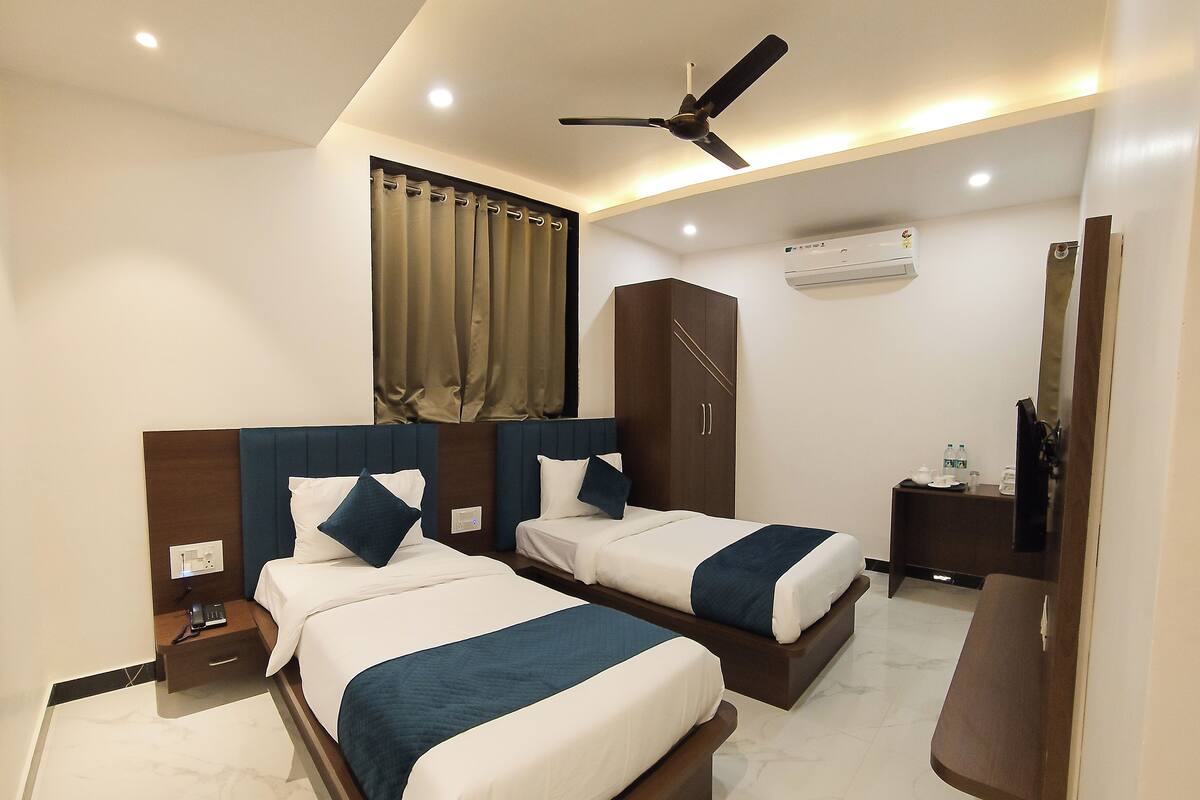 A hotel room with two beds, a ceiling fan, an air conditioning unit, a bedside table with a phone, and a small table with a bottle and glasses.