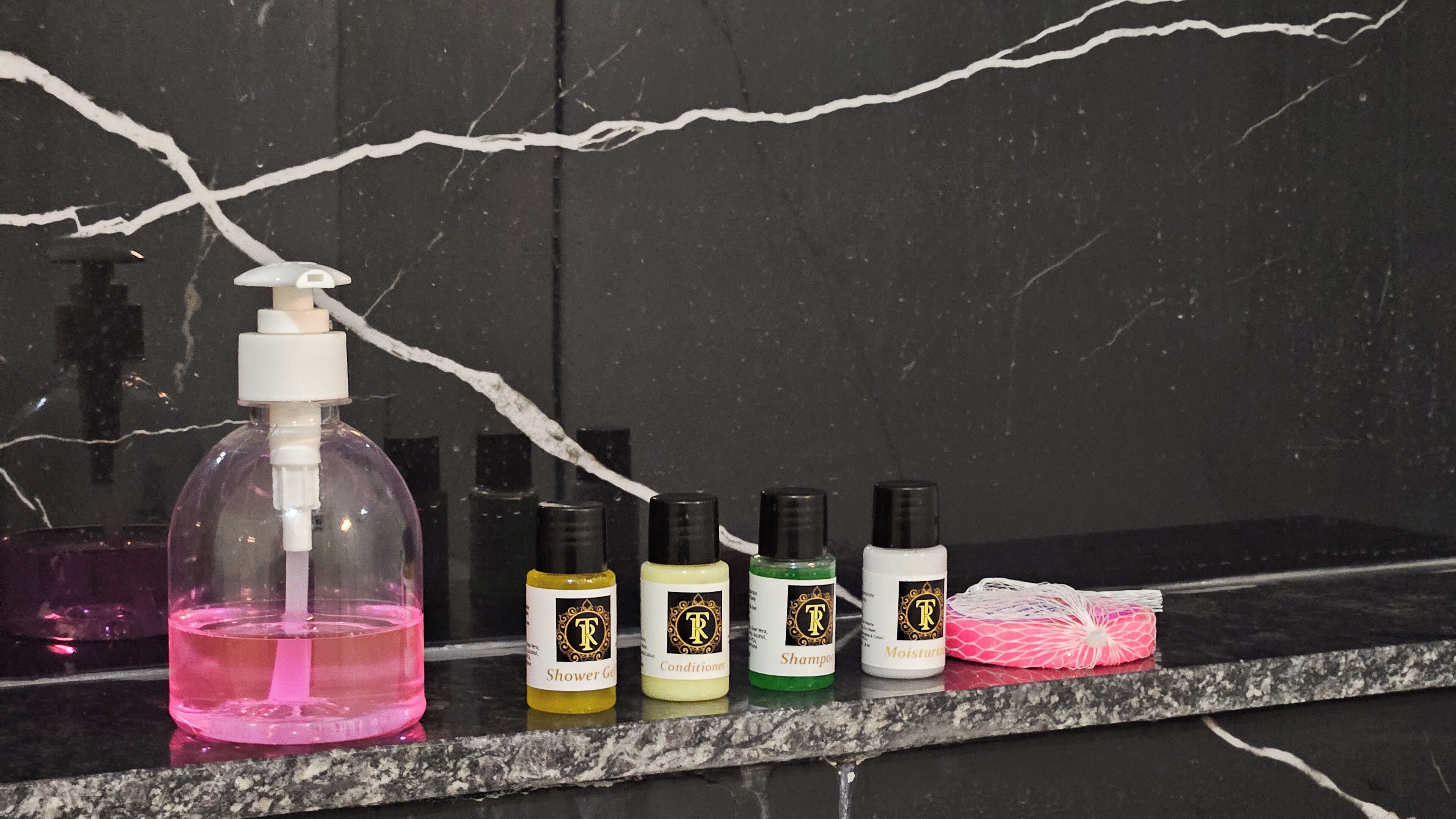 A bathroom counter with various toiletries including a pink bottle of liquid soap, four bottles of hair care products, and a pink hairbrush.
