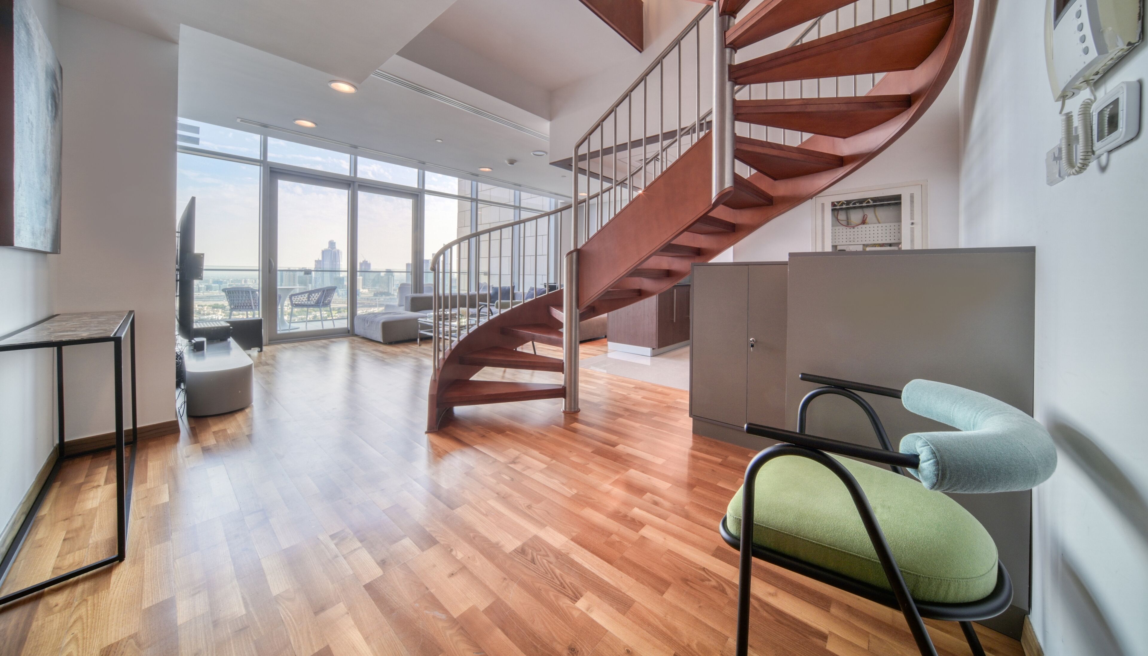 A spacious living area with a wooden floor, a modern staircase, and large windows offering a city view.
