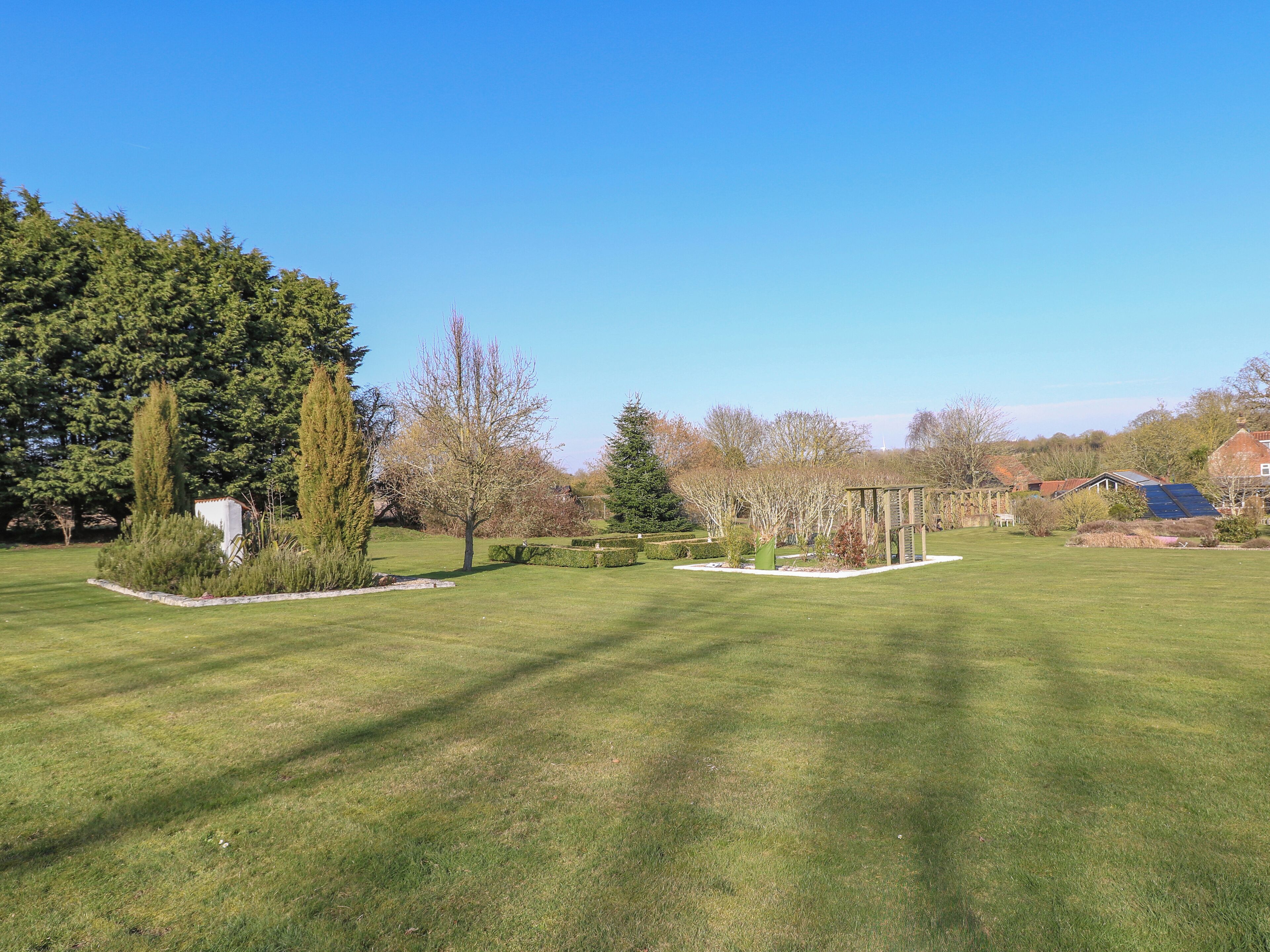 A well-maintained garden with a variety of trees and shrubs, a pergola structure, and a clear blue sky.