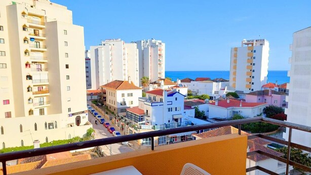 A balcony with a view of a coastal town, featuring multi-story buildings, a clear sky, and a road with parked cars.