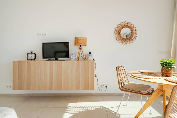 A modern living room with a TV on a wooden cabinet, a lamp, a clock, and a dining area with a round table and chairs.