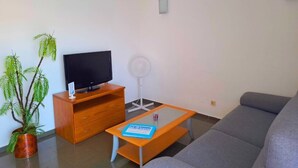 Una sala de estar con un sofá, un televisor sobre un mueble de madera, una mesita con un libro y una planta en maceta.