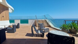 A sunlit outdoor deck with wicker furniture, a glass railing, and a view of the ocean.