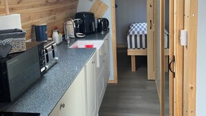 A compact kitchen with a built-in toaster, a coffee maker, and a microwave. There is a window above the sink and a small bed with a checkered bedspread in the background. - Starview by Mirriemora l Shetland l St Ninians Isle l Cosy Pod (Bigton)