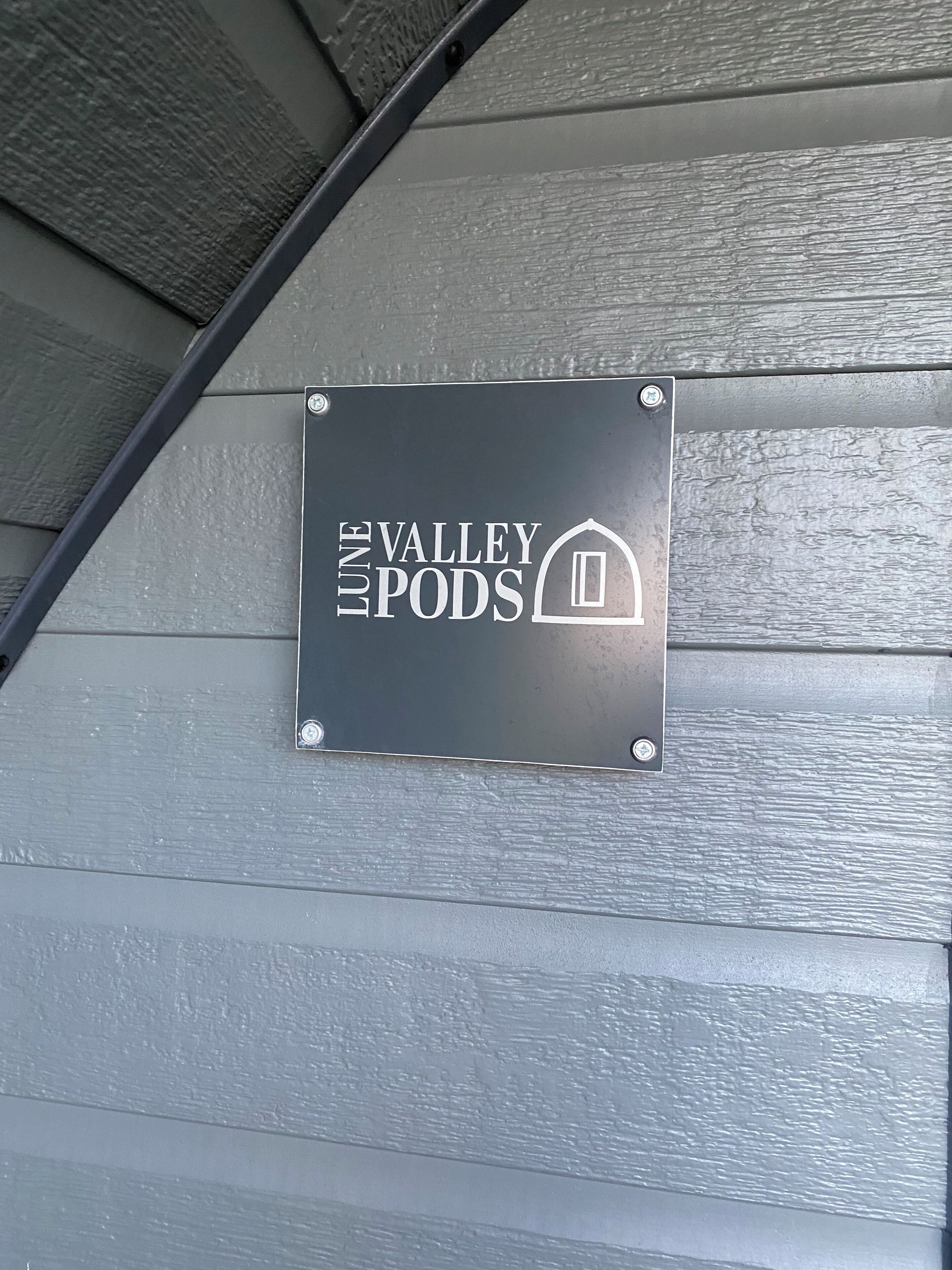 Une plaque métallique portant l’inscription « Valley Pods » et un symbole représentant une maison, fixée à un mur recouvert de bardage horizontal.