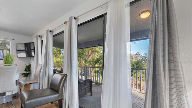 A modern room with a sliding glass door leading to an outdoor deck, a flat-screen TV mounted on the wall, and a seating area with a bench and chairs. - Bradenton Breeze  (Bradenton)