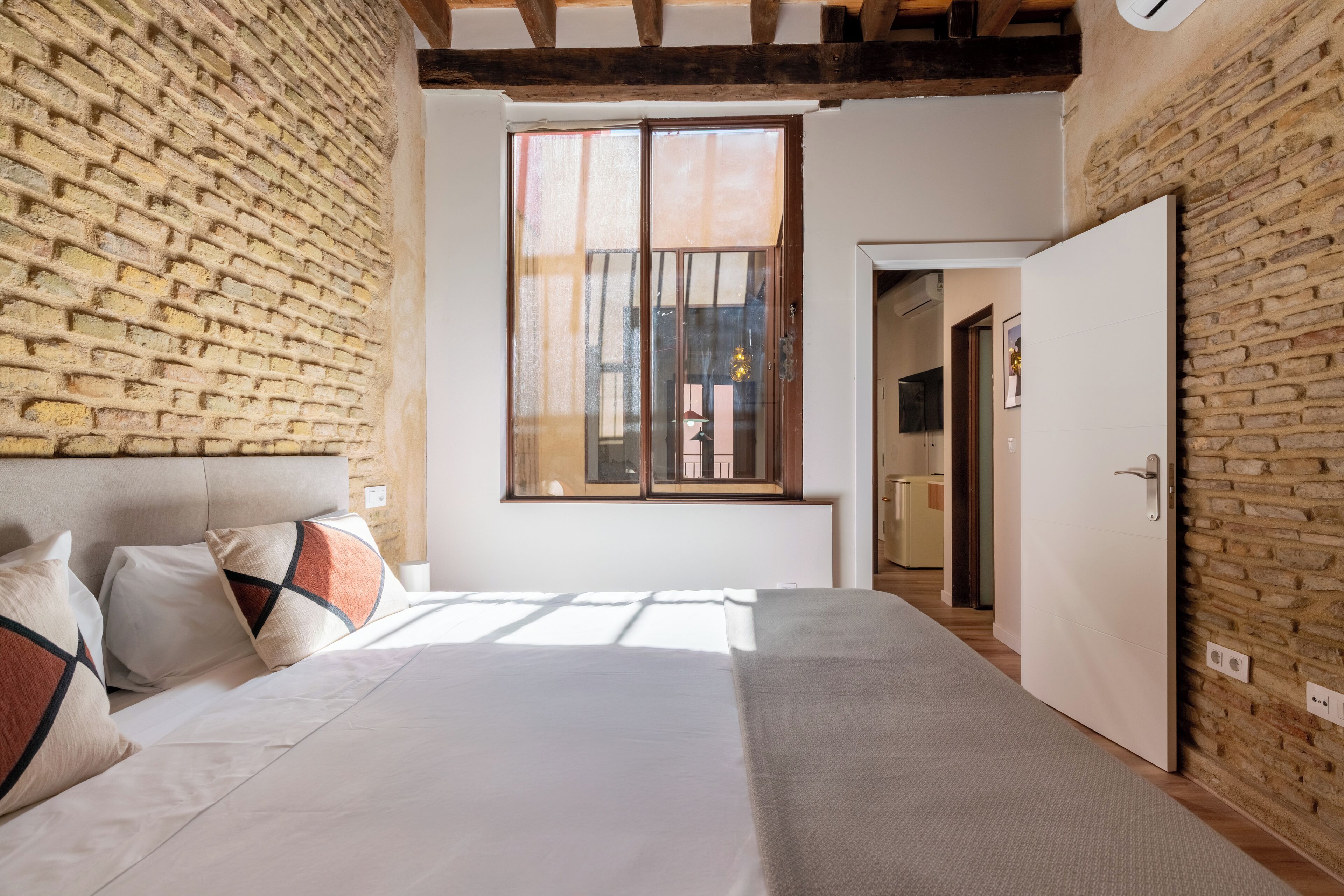 Quarto com cama grande, parede de tijolos, janela e porta que dá para outro cômodo.
