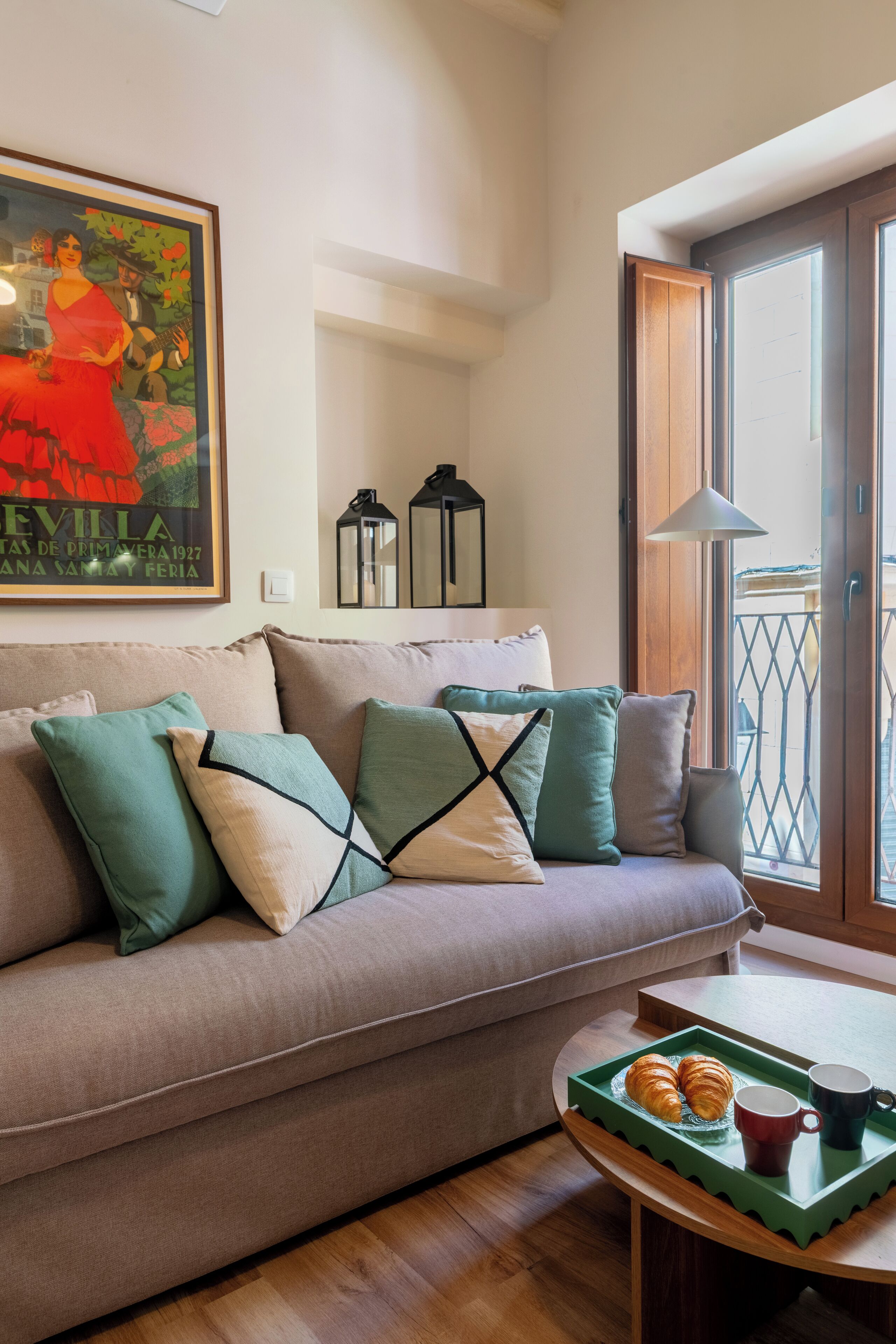 A living room with a sofa, pillows, a tray with croissants and mugs, a painting, and a balcony view.