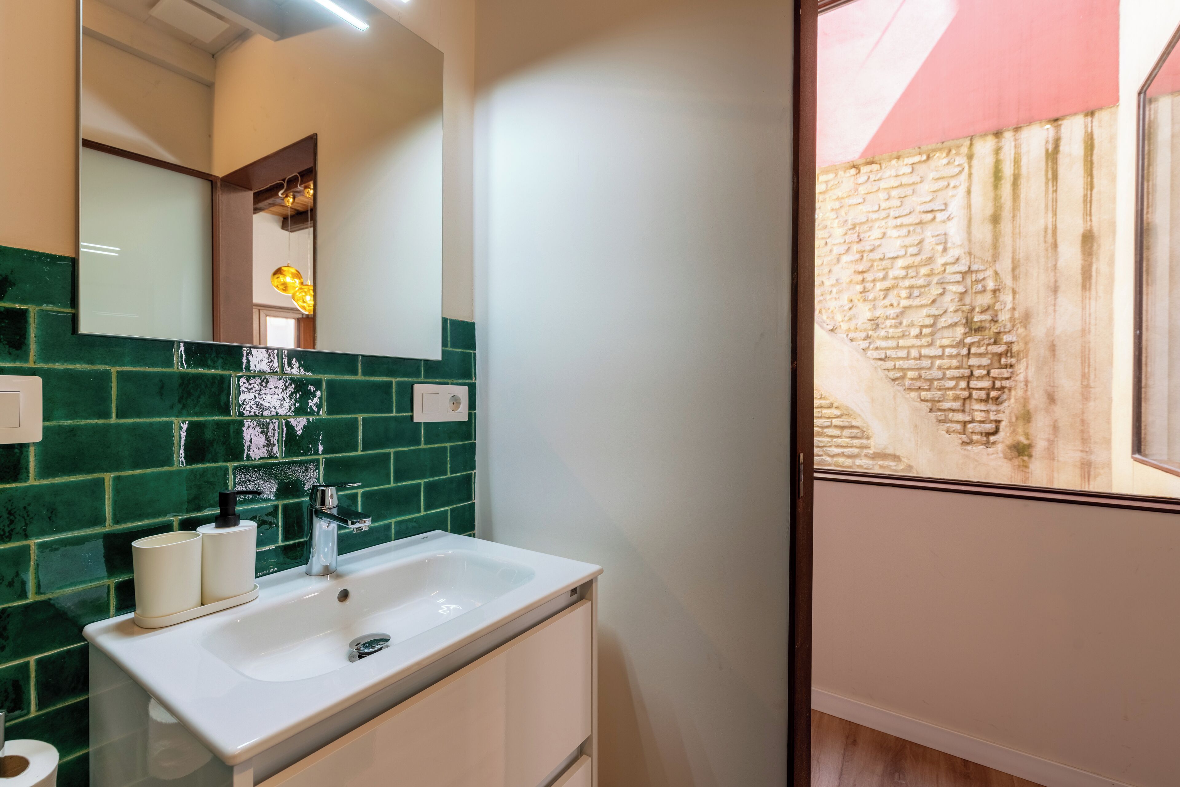 A bathroom with a white sink, green and white tiled backsplash, a mirror, and a window with a view of a brick wall.