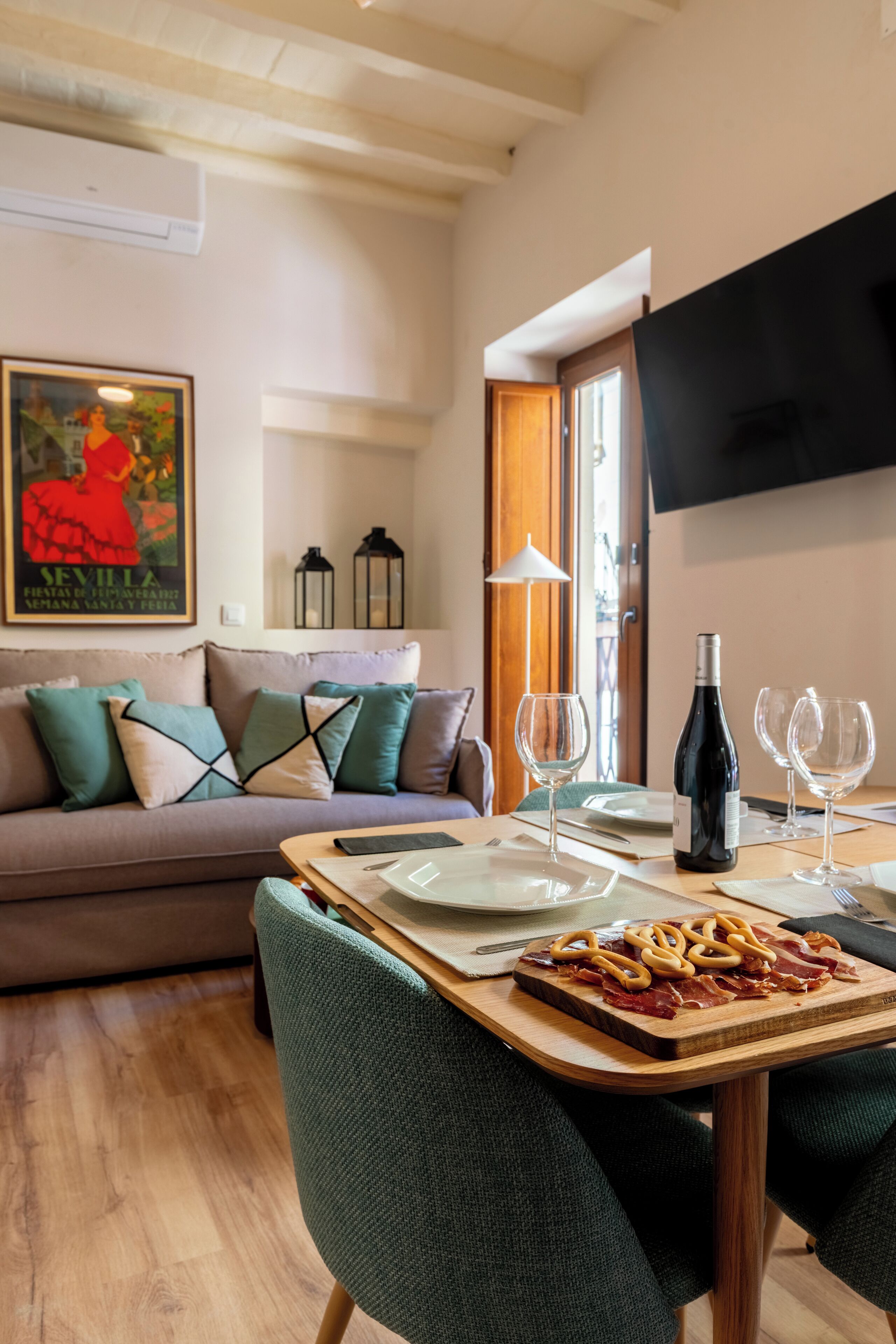 A dining area with a wooden table set for two, a bottle of wine, and a plate of food. There is a sofa with cushions in the background and a TV mounted on the wall.