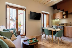 A modern apartment with a living room, dining area, and kitchen. There is a flat-screen TV mounted on the wall, wooden cabinets, and a dining table with chairs.