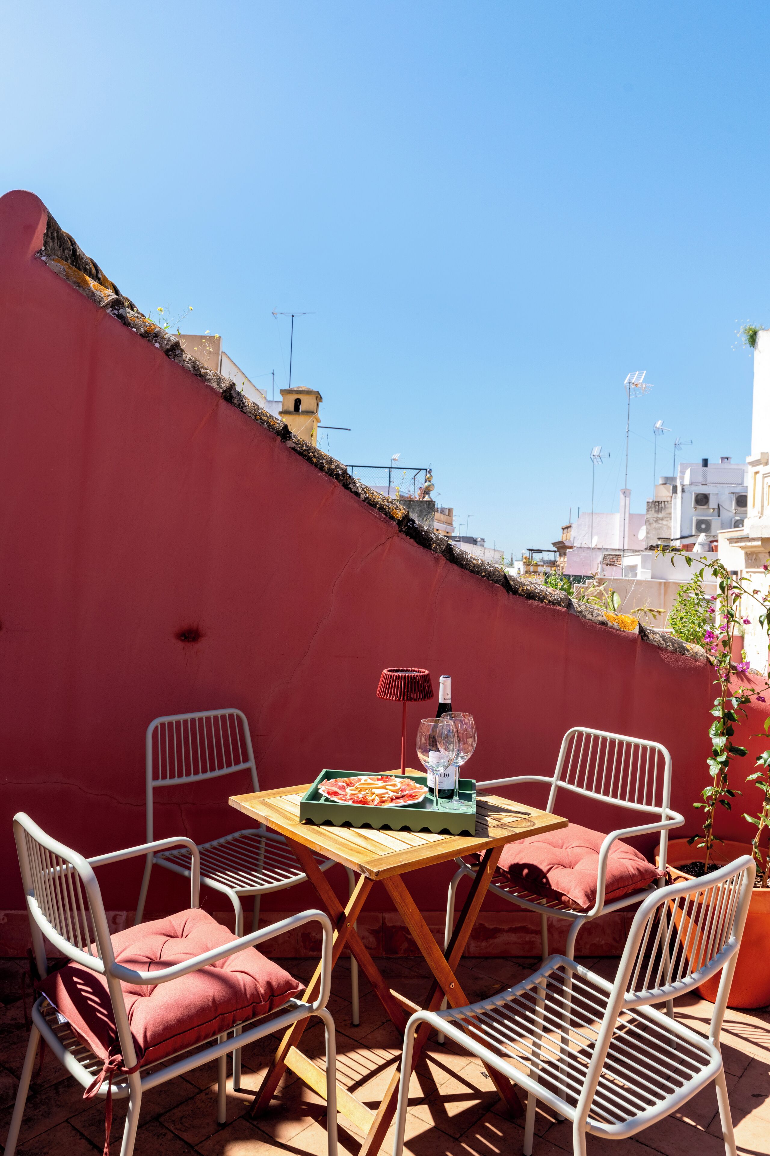 A rooftop terrace with a red wall, a wooden table, white metal chairs, and a bottle of wine.