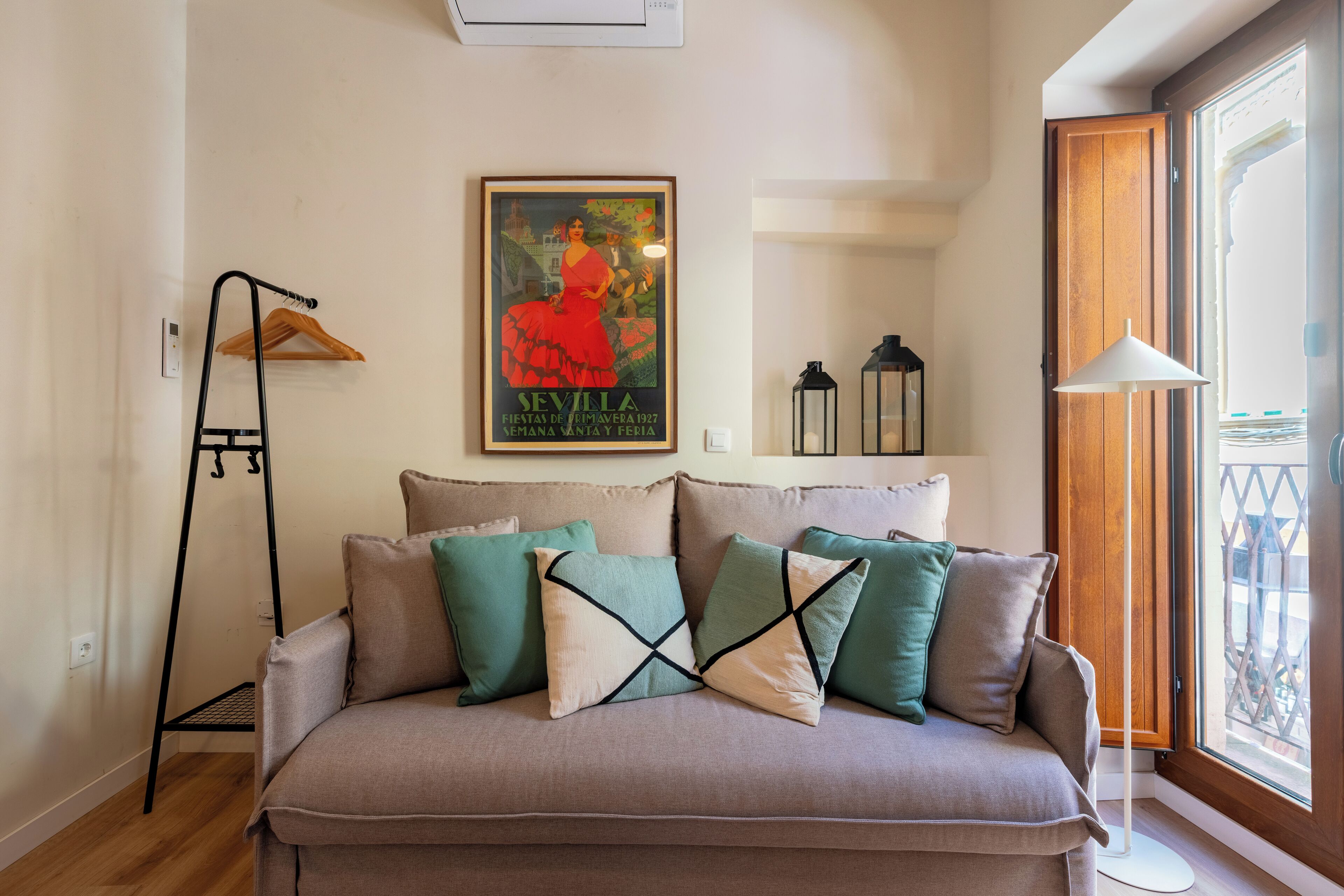 A living room with a beige sofa, green and white cushions, a wooden hanger, a framed picture, and a window with a view.