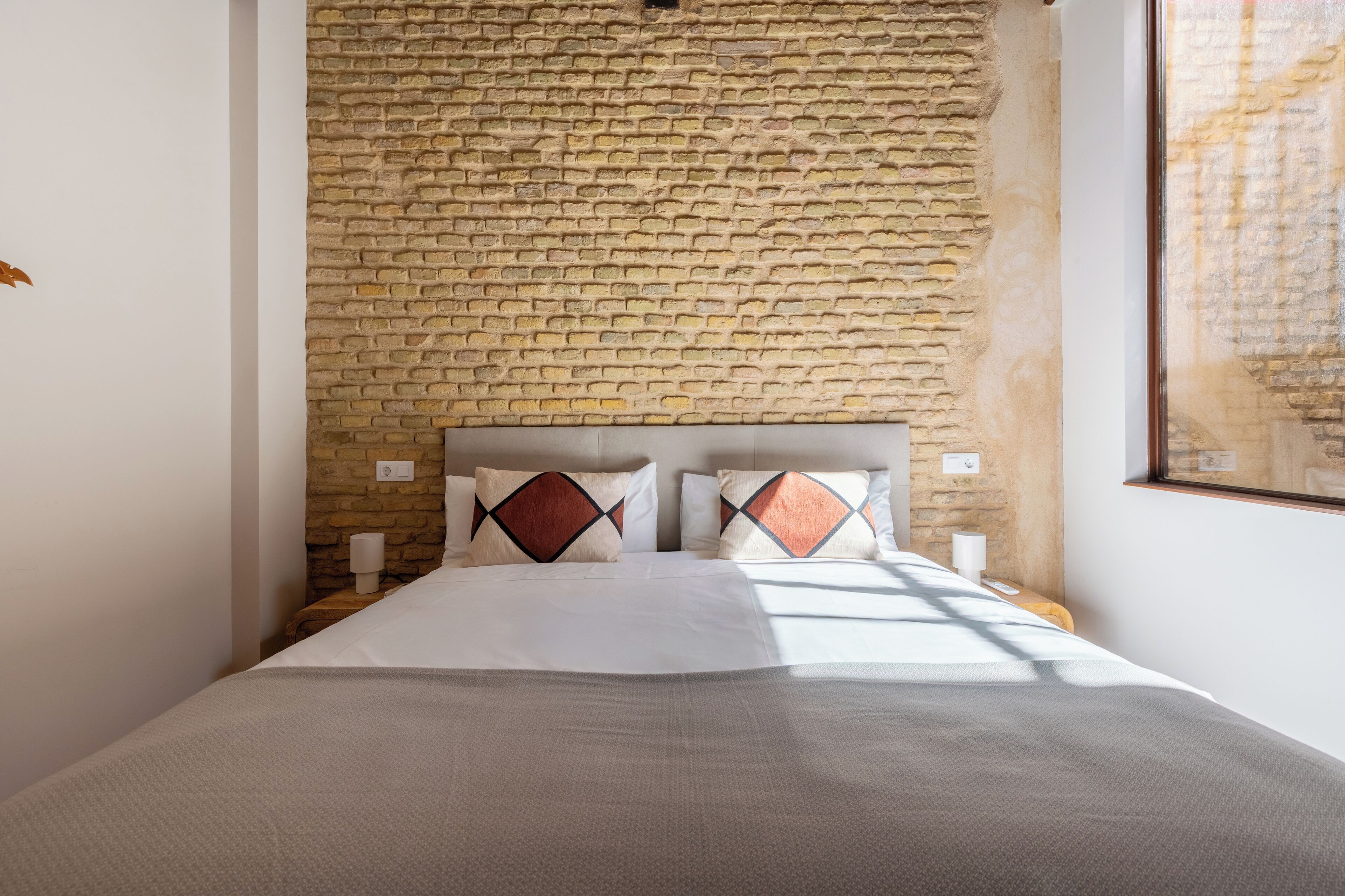 A bed with a headboard against a brick wall, two pillows, and a bedside table with a lamp.