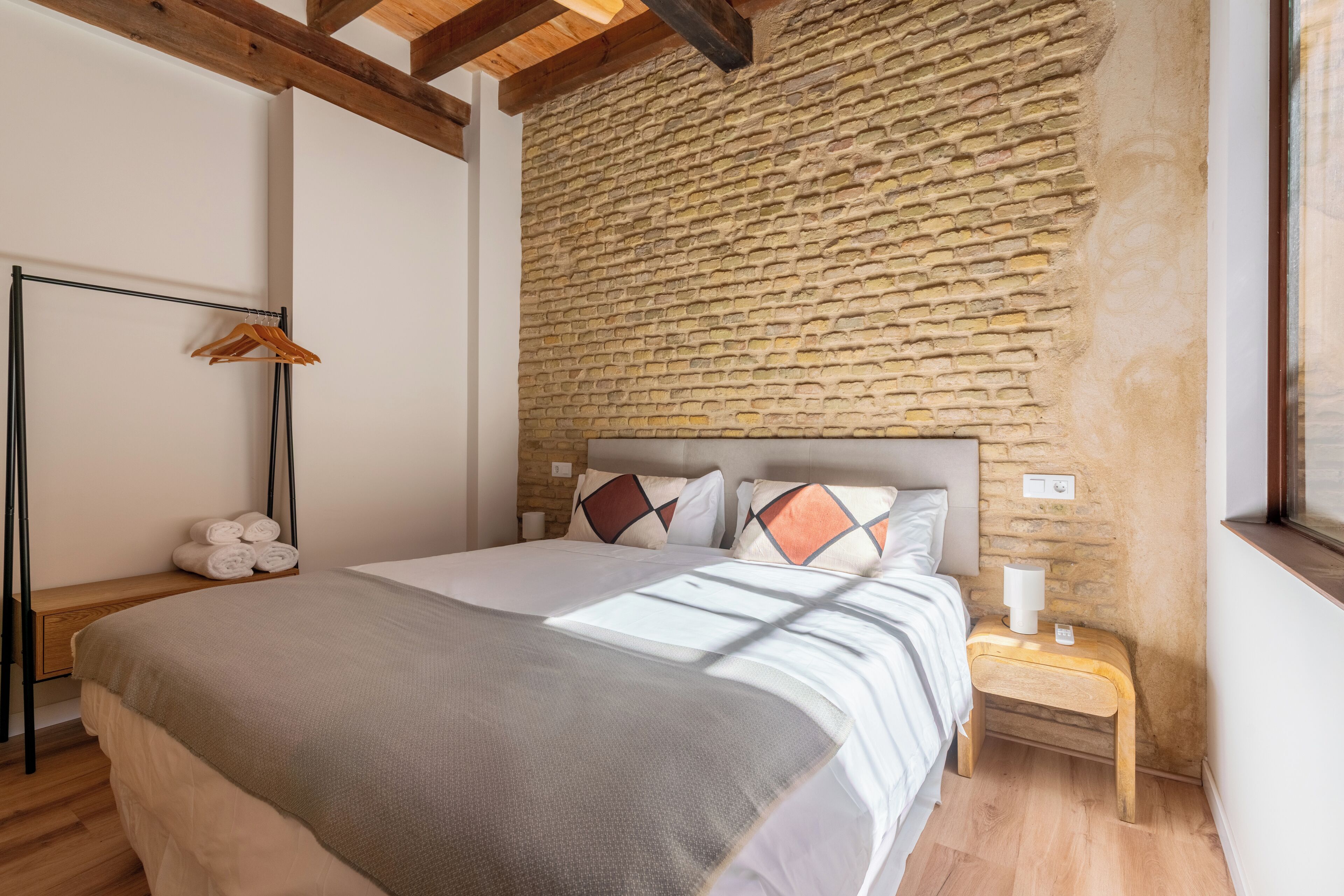 A bedroom with a stone wall, a wooden floor, a bed with a grey blanket, a bedside table with a lamp, and a wooden bench.