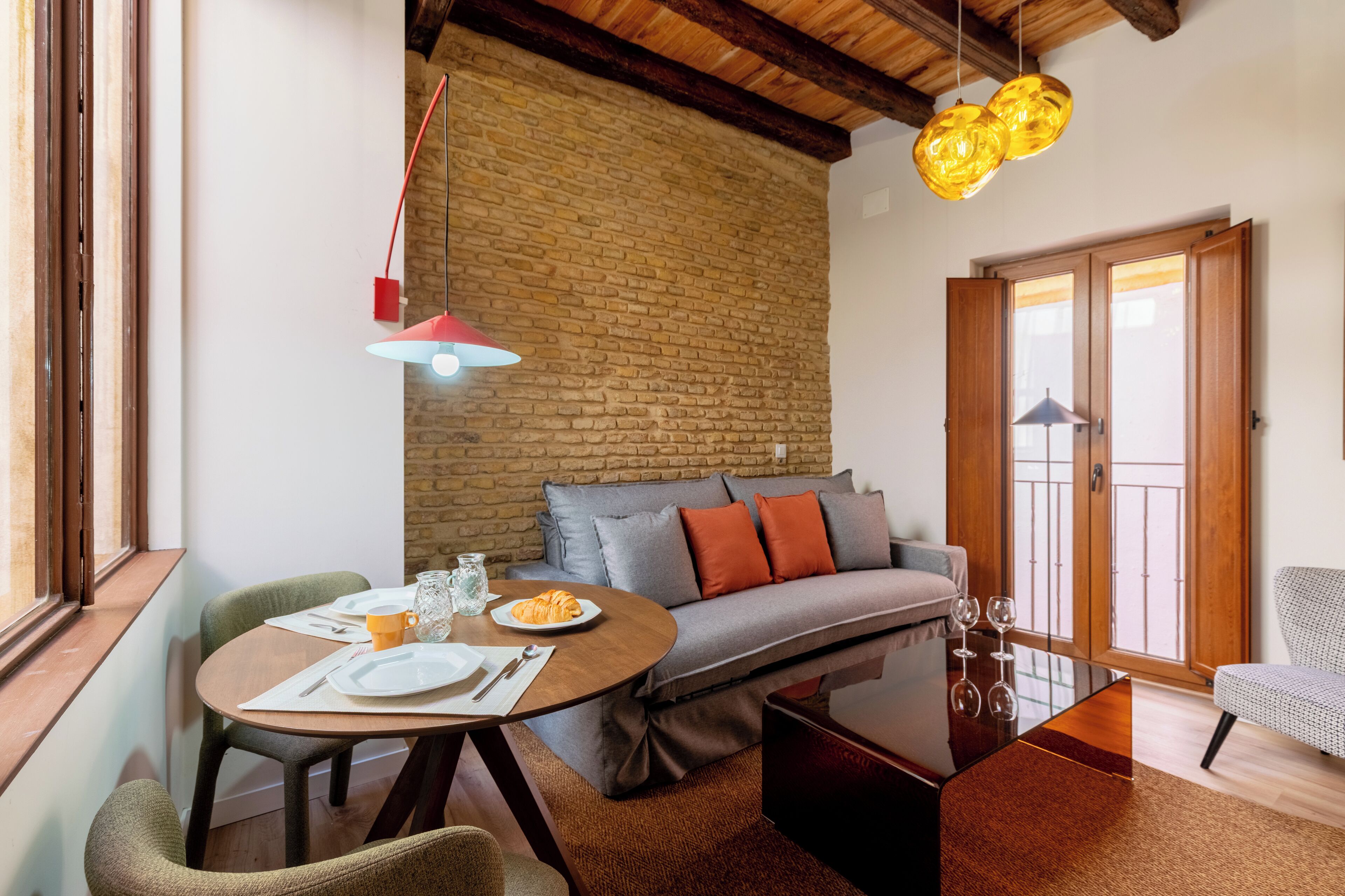 Ein gemütlicher Wohnbereich mit Backsteinwand, grauem Sofa mit orangen Kissen, Couchtisch und Esstisch mit Stühlen.