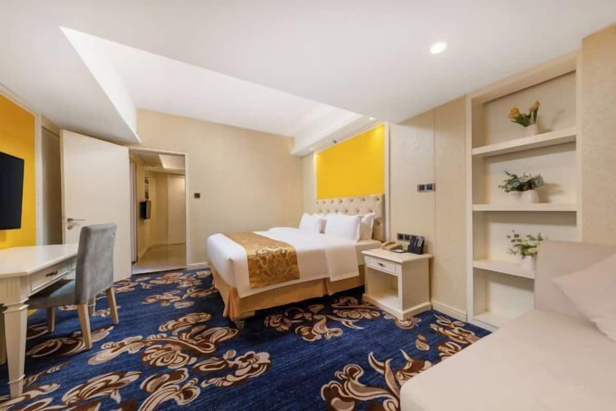 Una habitación de hotel con cama, mesita de noche, silla, escritorio y estanterías con objetos decorativos.