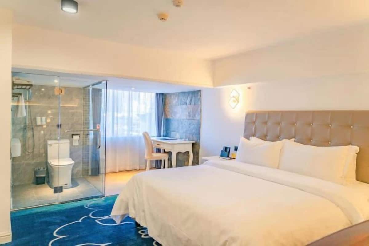 Habitación de hotel con cama, escritorio, silla, cabina de ducha de vidrio y vistas al exterior.