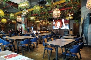 En restauranginteriör med bord, stolar, hängande växter och en tv-skärm.