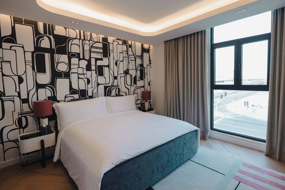 Ein modernes Hotelzimmer mit einem großen Bett, einer Wand mit Muster, einem Nachttisch mit Lampe und Blick auf die Stadt durch ein großes Fenster.