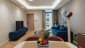 A modern living room with a flat-screen TV, a dining table with a fruit bowl, a blue sofa, and a large window with curtains. - The Muse Hotel  (Lusail)