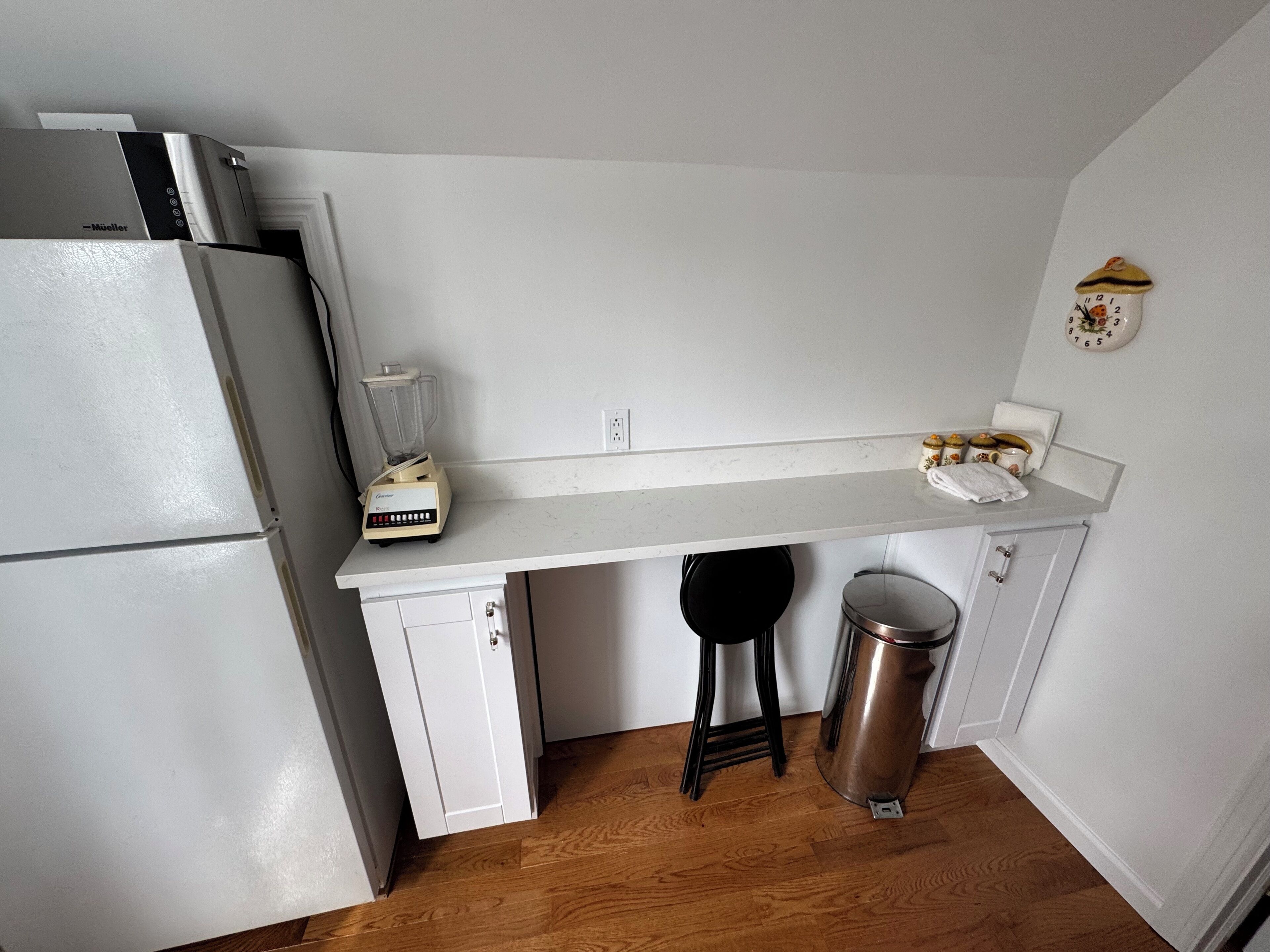 A compact kitchen with a white countertop, a blender, a stainless steel trash can, and a refrigerator.
