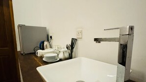 A bathroom with a white sink, a mirror, and a towel rack. There is a door on the left and a small table with a kettle and cups in the background. - Crips Hill Apartments (Galle)