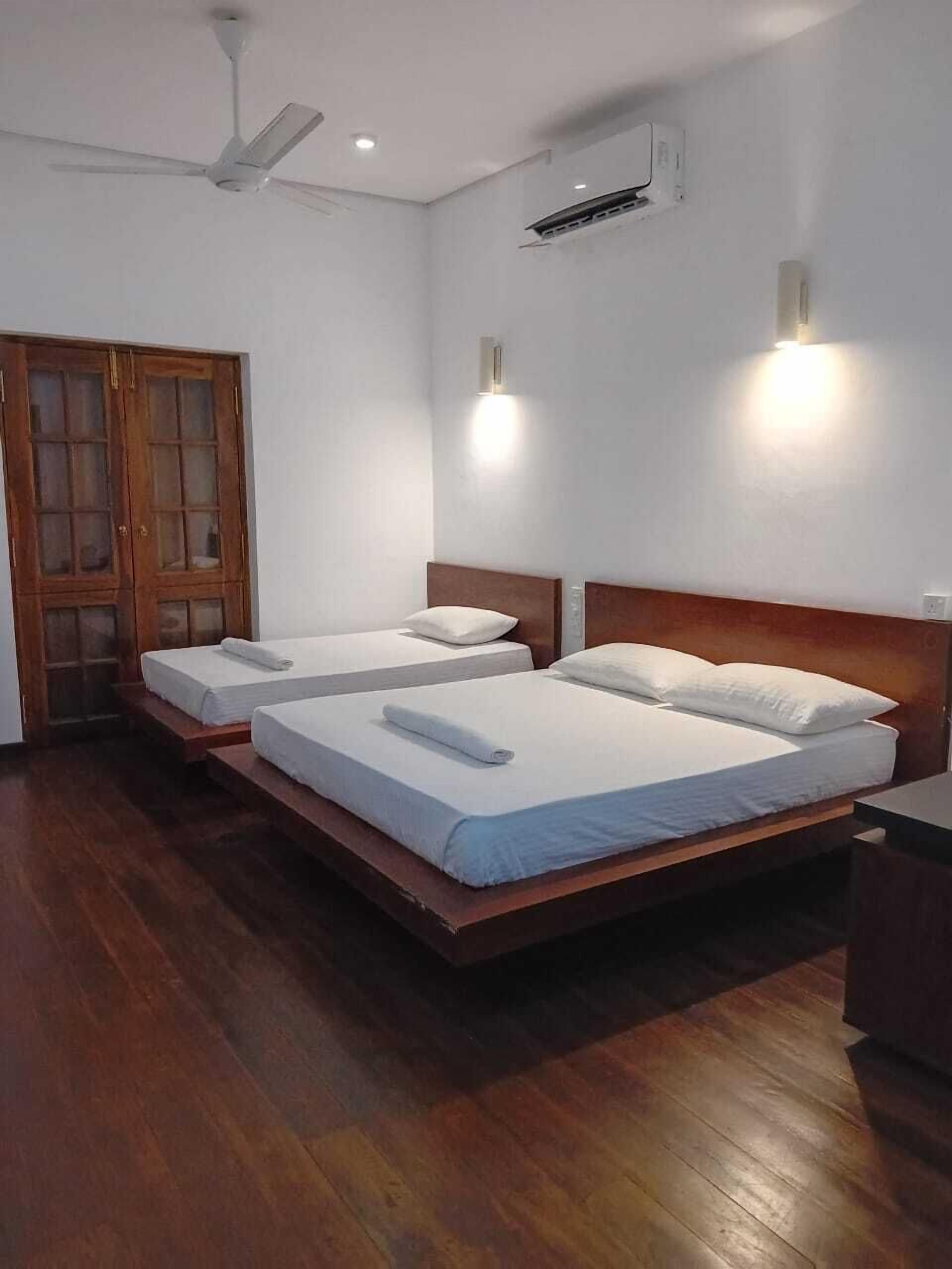 Habitación con dos camas, piso de madera, ventilador de techo e iluminación en la pared.