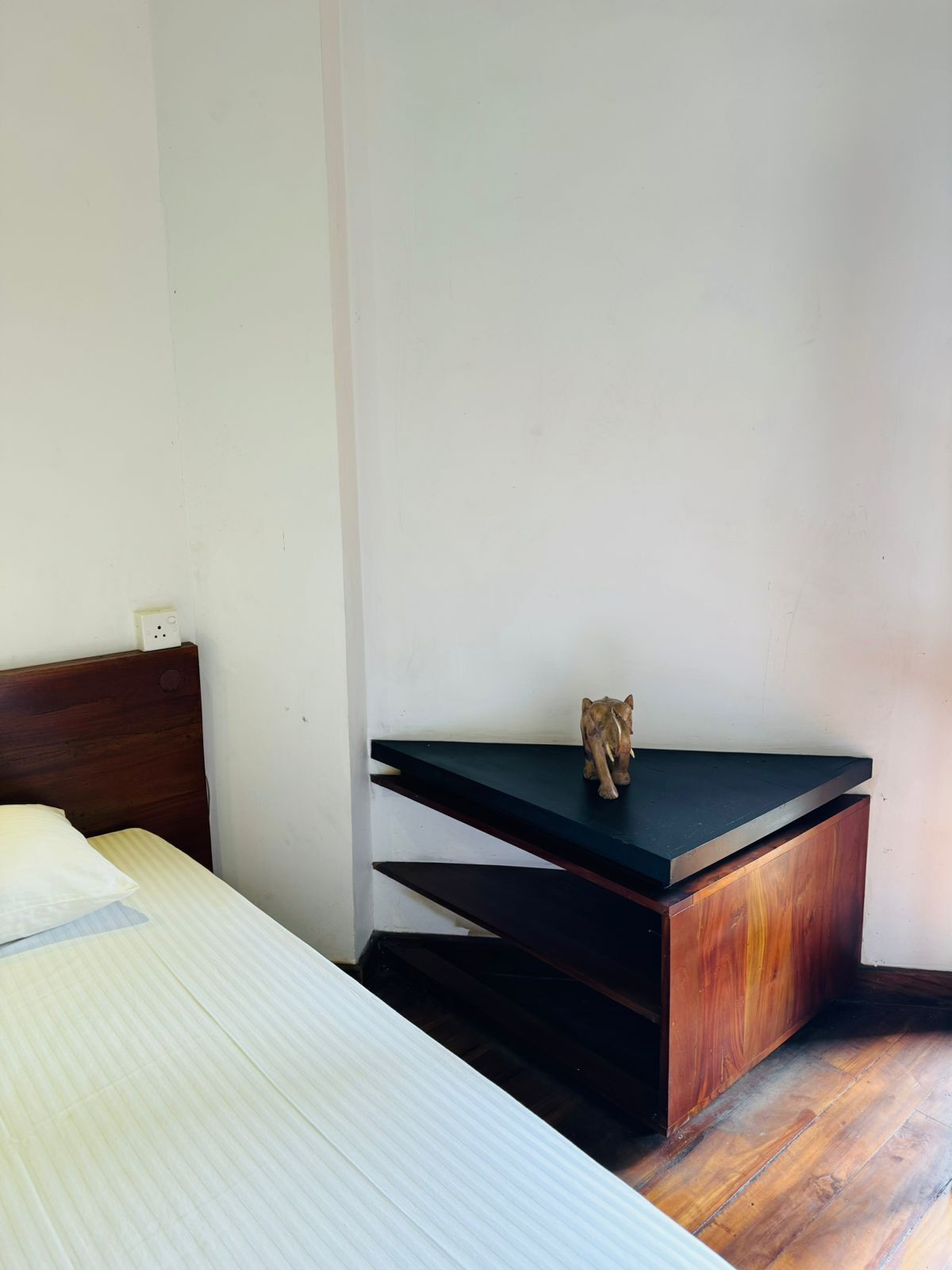 Un dormitorio con cama, cabecera de madera, mesita de noche con una pequeña figurita de elefante y un escritorio de color oscuro.