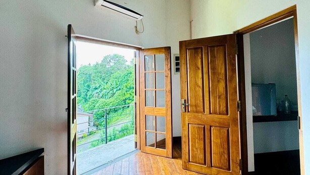 A room with wooden flooring, a large wooden door, and a sliding glass door leading to a balcony with a view of greenery. - Crips Hill Apartments (Galle)