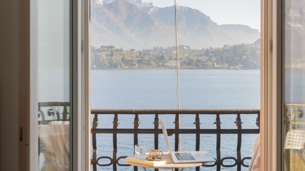 A balcony with a laptop, a glass of water, and a book on a small table, overlooking a lake and mountains. - Cadenabbia Lake Side Apartment 3 (Cadenabbia)