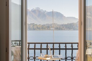 Un balcón con una computadora portátil, un vaso de agua y un libro sobre una mesita, con vistas a un lago y montañas.