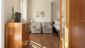 A bedroom with a wooden dresser, two armchairs, a bed, and a painting on the wall.
