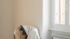 En hörna i ett rum med en flätstol täckt av ett vitt tyg med svarta mönster, ett vitt element och ett fönster med utsikt över byggnader.