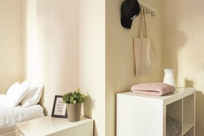Une chambre à coucher bien rangée, avec un lit, une table de chevet ornée d’un cadre contenant une citation, une plante en pot, une commode blanche, une lampe blanche et un porte-chapeaux et porte-sacs fixé au mur.