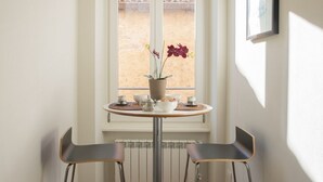 A dining area with a table set for two, a potted plant, and a window with a view of a tiled roof.