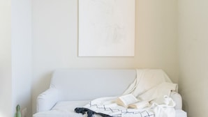 A white sofa with a blanket and a book, a framed artwork above it, and a small plant on the floor.