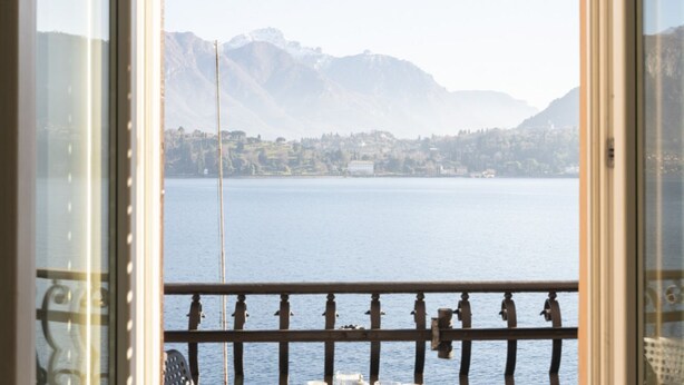 A view from a window with a balcony overlooking a lake and mountains.