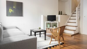 Ein modernes Wohnzimmer mit einem grauen Sofa, einem schwarzen Couchtisch, einem Korbstuhl, einem Fernseher auf einem weißen Regal und einer Treppe.