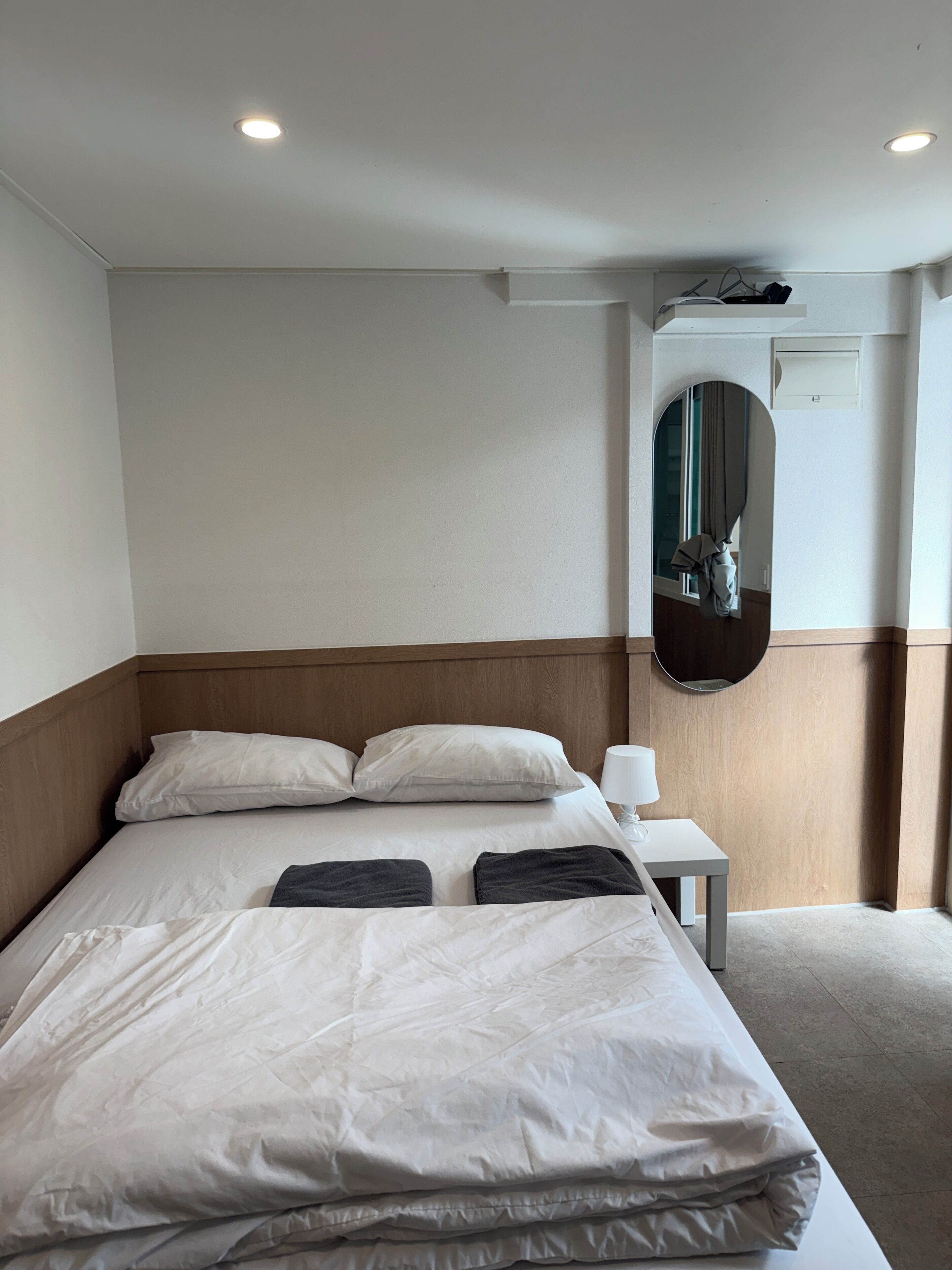 A hotel room with a bed, a bedside table with a lamp, an air conditioning unit, and a mirror on the wall.