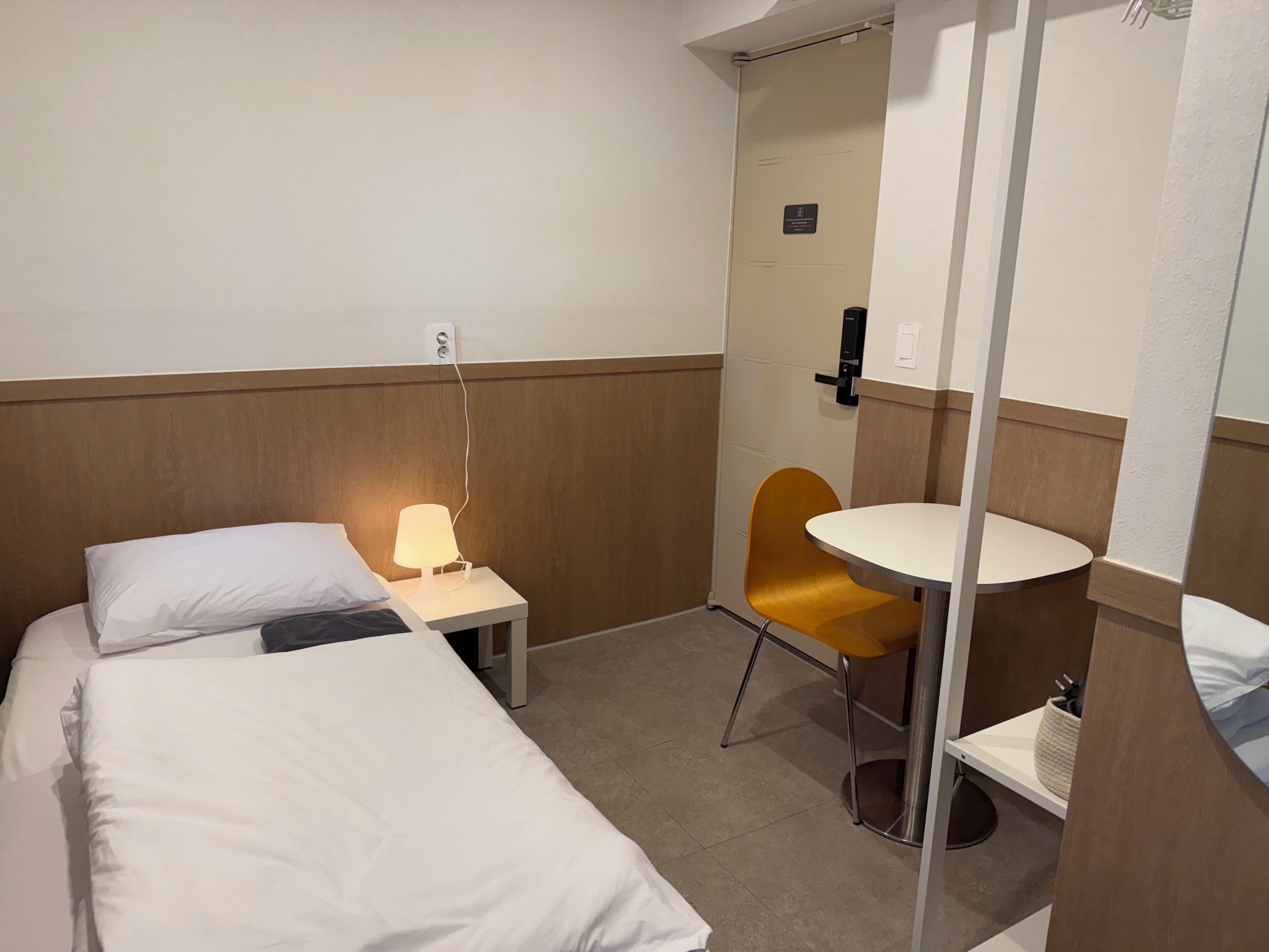 A hotel room with a bed, a bedside lamp, a small table, a chair, and a mirror.