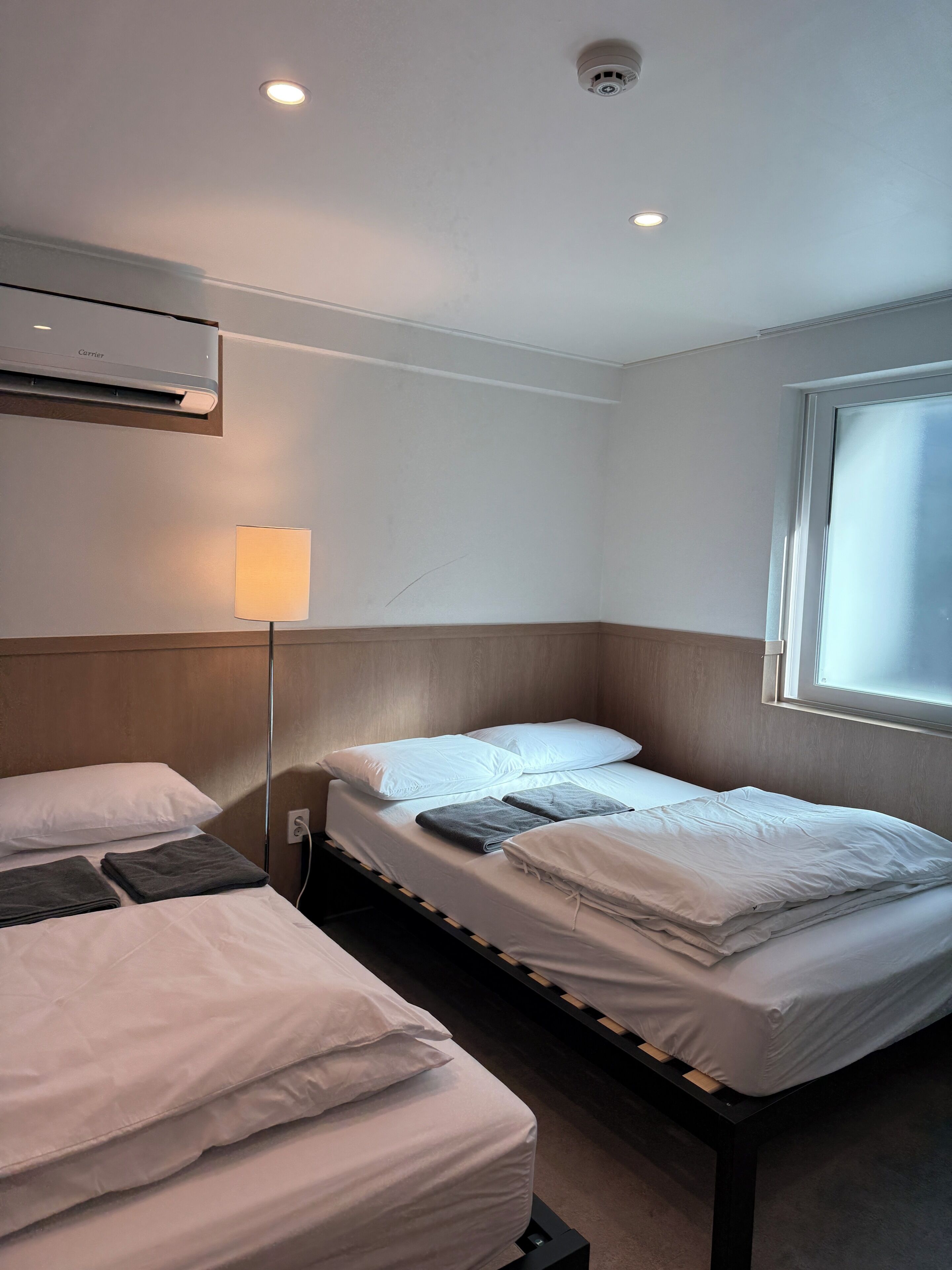 A hotel room with two beds, a window, an air conditioner, and a lamp.