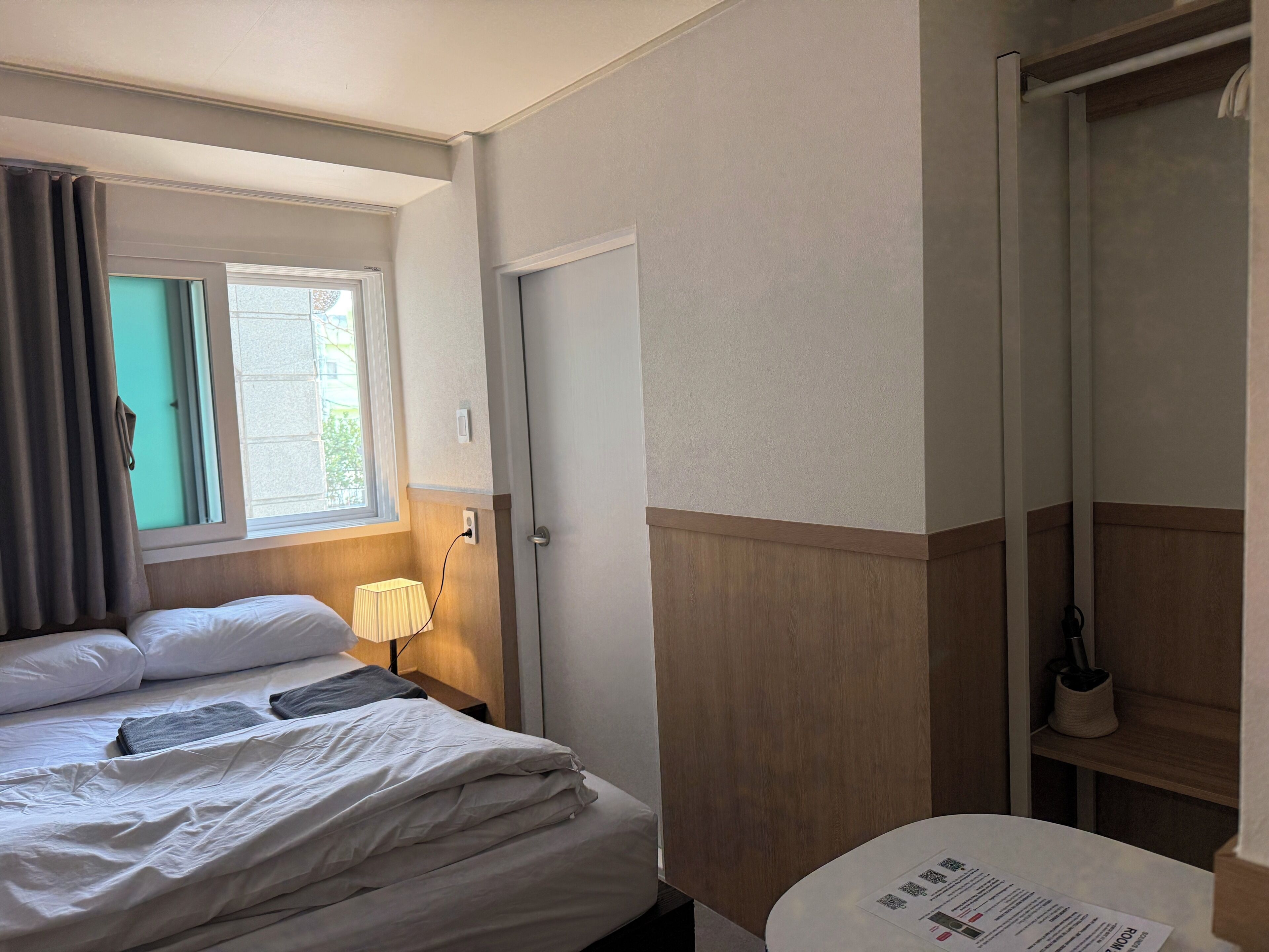 A hotel room with a bed, a bedside table with a lamp, a window with curtains, a door, and a small round table with a menu.