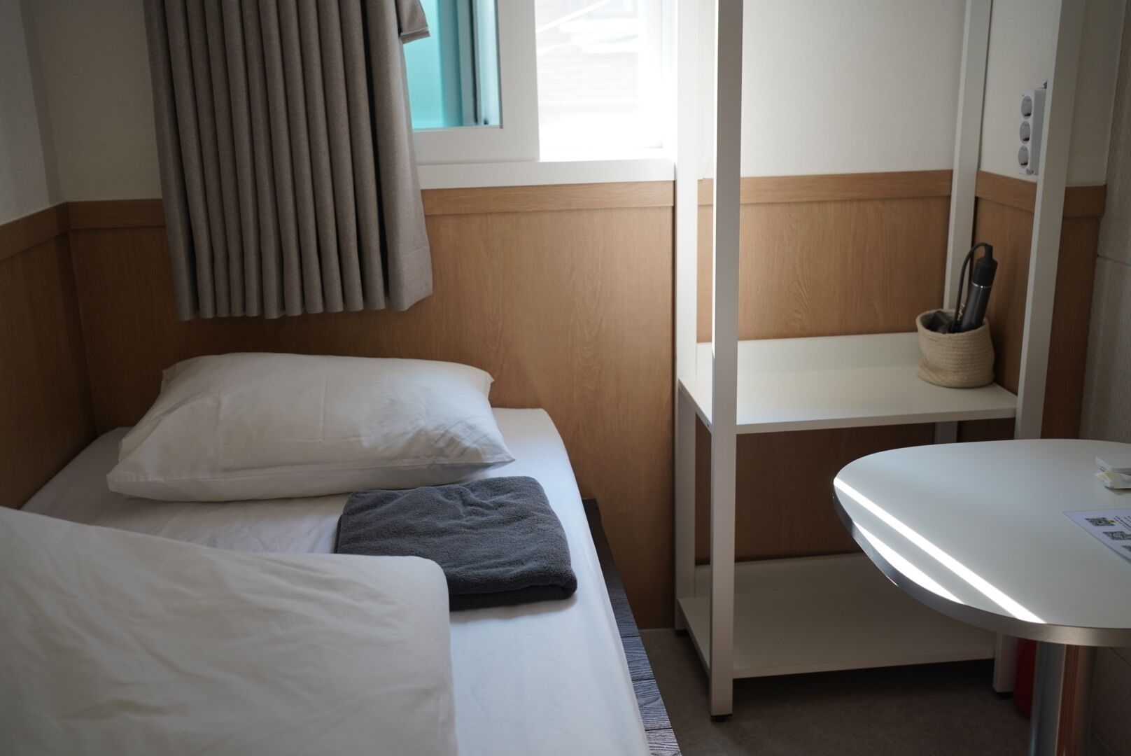 Una habitación de hotel pequeña con cama, mesita de noche, una silla, ventana con cortinas y una estantería en la pared.