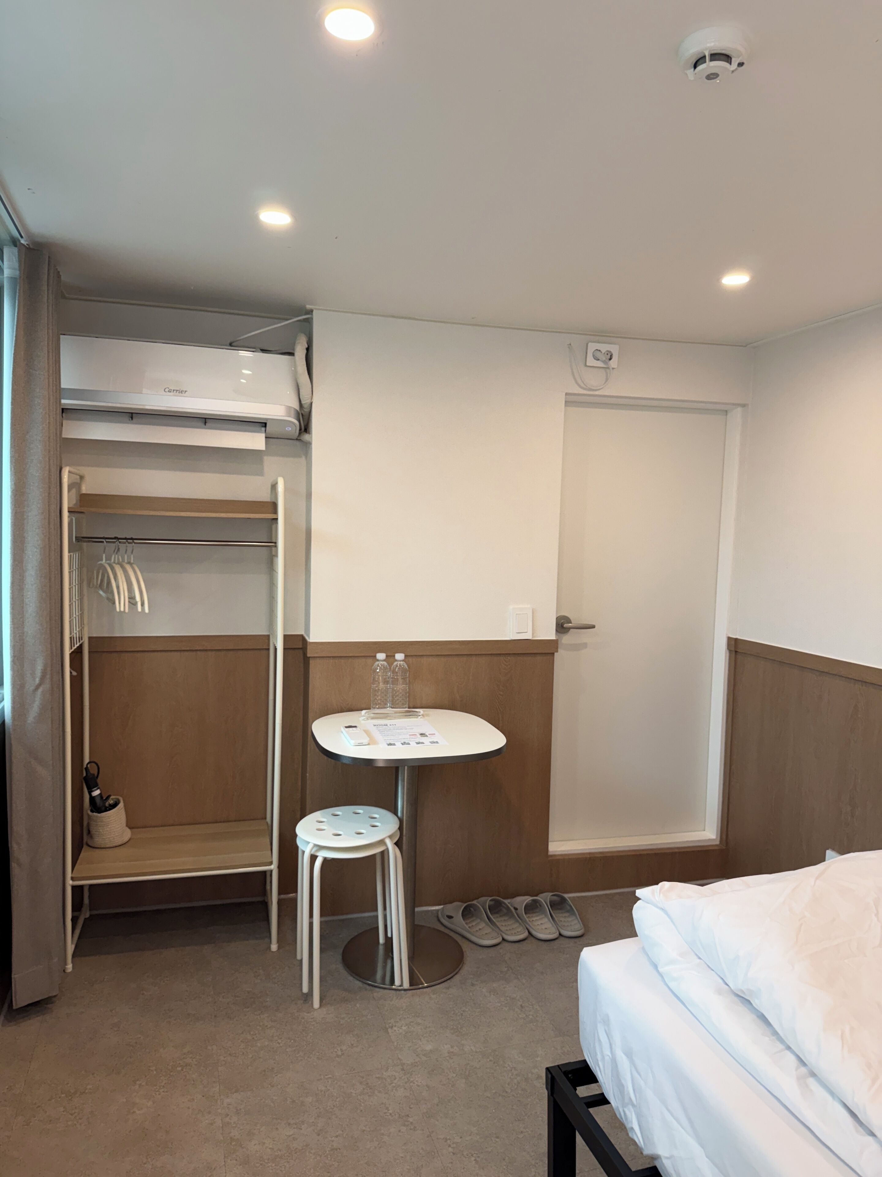 A hotel room with a bed, a small table with a chair, a closet with hanging clothes, and a shower area.