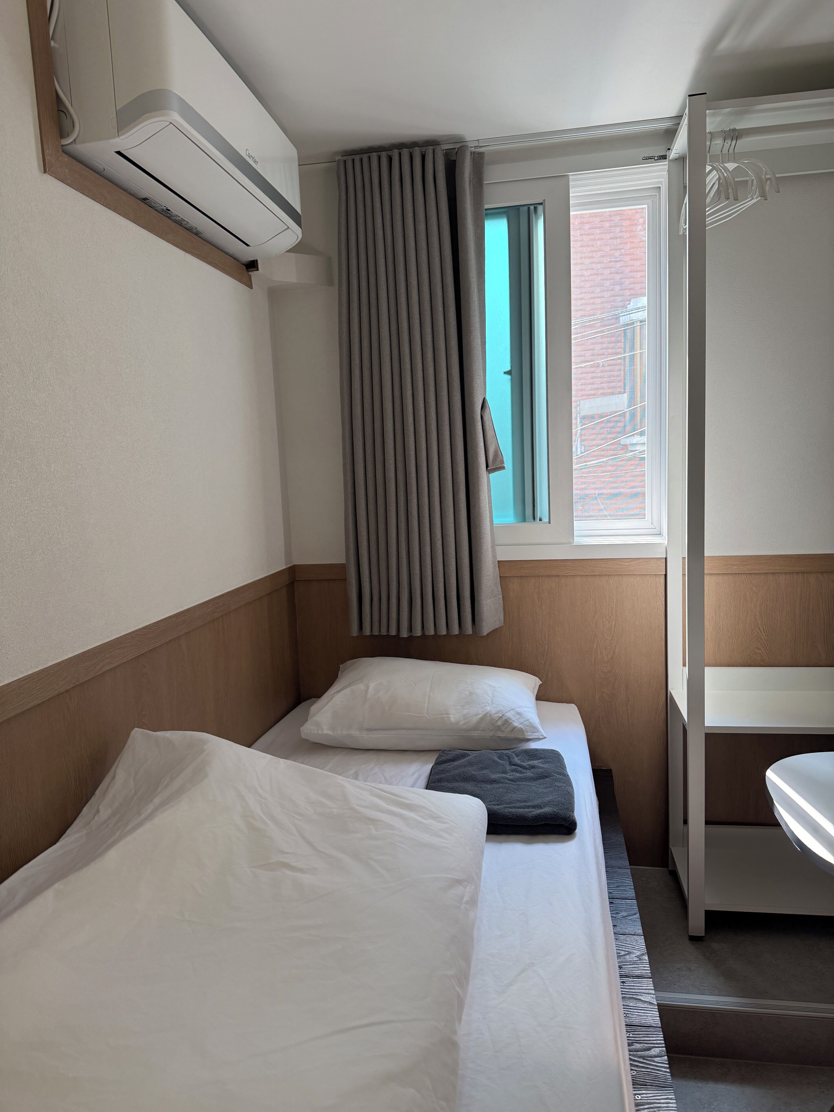 A hotel room with a bed, a window with curtains, an air conditioning unit, and a bathroom with a sink.
